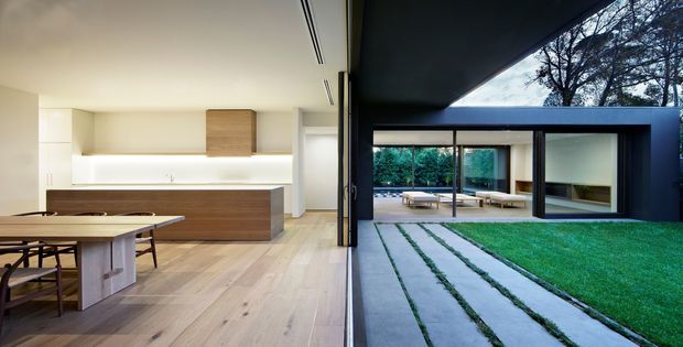 Open-plan contemporary kitchen and dining area opening directly onto a landscaped courtyard. The interior has pale timber flooring, white walls and minimalist built-in cabinetry with a dark wood island, while expansive black-framed glazing creates a strong indoor–outdoor connection. The courtyard features a lawn, linear concrete or stone stepping strips, mature trees and a covered outdoor seating area.