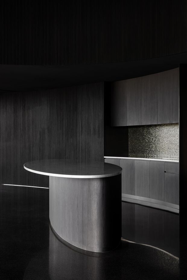 A dramatic, monochromatic contemporary kitchen with a curved dark island, integrated cabinetry, a textured metallic mosaic splashback, and vertically ribbed wall surfaces. The space has a highly refined, sculptural appearance with glossy dark flooring and concealed lighting.