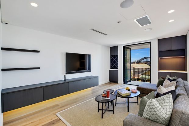 Contemporary living room with a large grey sectional sofa, black nesting coffee tables, a wall-mounted television, low dark built-in media cabinetry, open shelving, and a light timber floor. The room has white walls and ceiling with recessed lighting, a glazed door opening to a balcony, and dark built-in joinery with warm integrated shelf lighting.