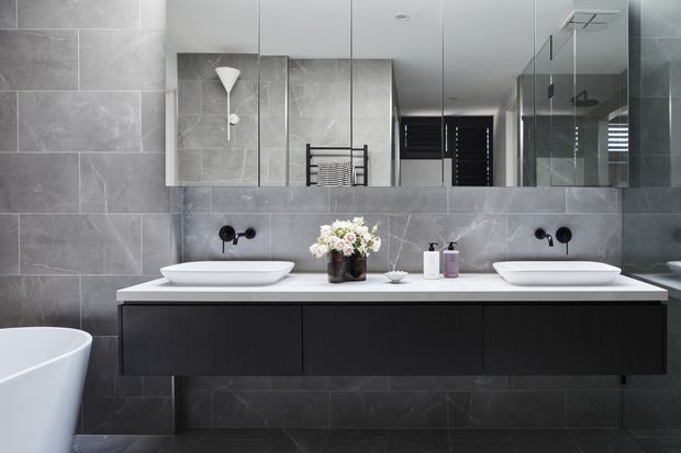 Contemporary bathroom featuring a double floating vanity with white vessel basins, dark cabinetry, a white benchtop, grey large-format wall and floor tiles, wall-mounted black tapware, and a large mirrored wall. A freestanding bathtub is partially visible at left, with a glass-enclosed shower reflected at right.
