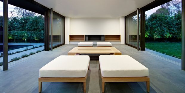A minimalist, open-plan living room with expansive floor-to-ceiling corner glazing opening to landscaped gardens and a pool terrace. Two low upholstered lounge benches face a timber coffee table and a linear built-in fireplace and shelving unit. The room has a pale smooth concrete floor, white walls and ceiling, dark-framed glazing, and restrained timber joinery.