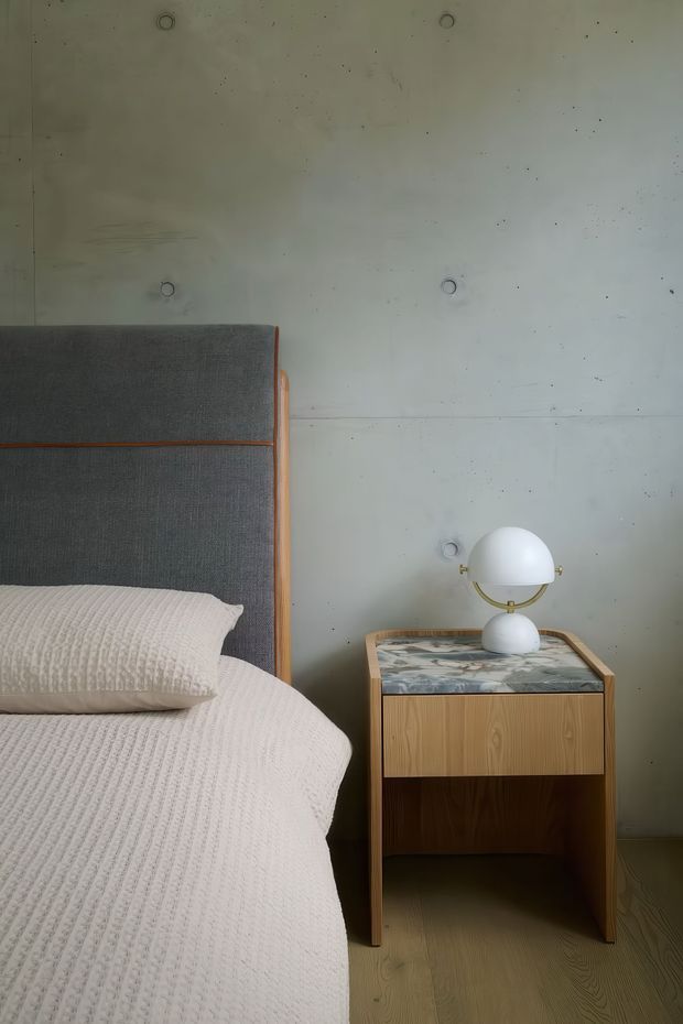 A close-up of a calm, contemporary bedroom corner featuring a partially visible bed with a tall charcoal-grey upholstered headboard edged in timber, white textured bedding, and a compact timber bedside table with a patterned stone top. A spherical white and brass table lamp sits on the nightstand. The background wall is raw pale grey concrete with visible formwork joints and circular tie marks, while the floor appears to be light timber.