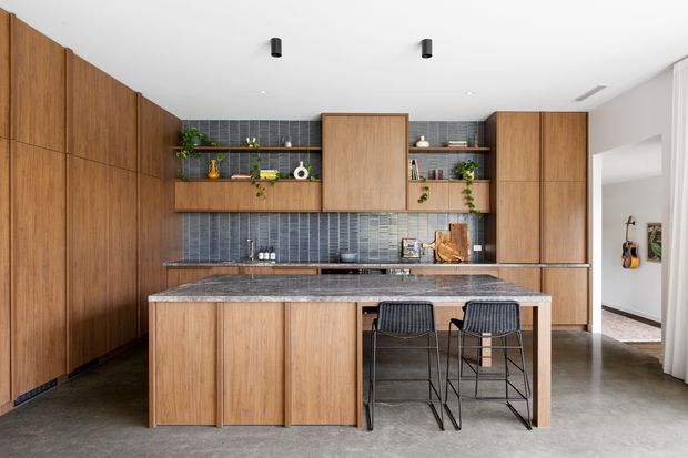 Contemporary kitchen with extensive warm timber cabinetry, a central island with two black bar stools, dark blue-grey vertically textured tiled splashback, and open display shelving with plants and kitchenware. The space has polished concrete flooring, a white ceiling with recessed and black cylindrical lighting, and a clean, refined fit-out.