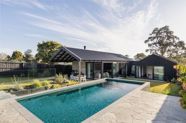 A contemporary single-storey residence surrounding a landscaped swimming pool. The home has dark vertical cladding, a pitched metal roof with exposed triangular gable framing, extensive glazed doors, and a broad pale stone-paved pool terrace. Lawn, planting, fencing, and mature trees surround the property.