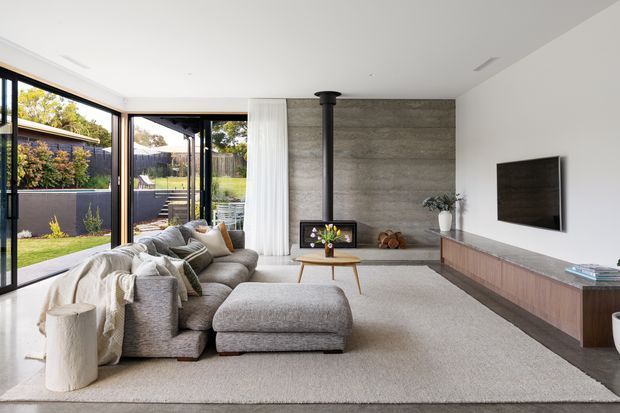 A bright contemporary living room with full-height glazed sliding doors opening to a landscaped outdoor area. The room features a large light-grey sectional sofa and ottoman, pale carpet, a textured grey concrete-look feature wall with a central black freestanding fireplace, a timber-toned built-in media console, wall-mounted television, and sheer white curtains. Minimal styling and abundant natural light create a calm, refined atmosphere.