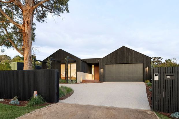 Street-level exterior view of a contemporary single-storey residence with a dark vertically clad facade, multiple gabled roof forms, a large integrated garage, and a recessed glazed entry. A pale concrete driveway leads through a dark slatted boundary fence and gate, with established eucalyptus trees and restrained planting around the property.