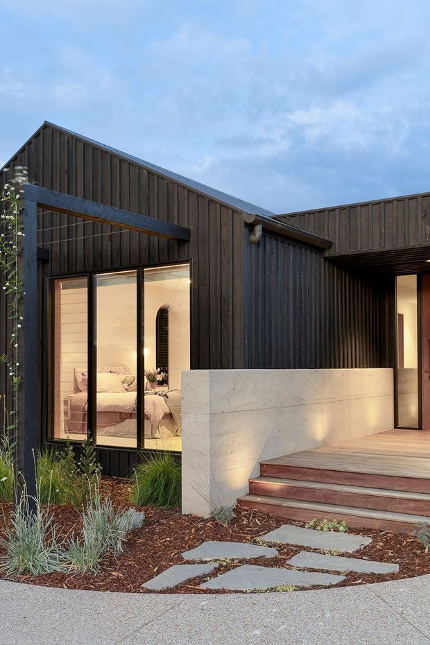 Exterior view of a contemporary single-storey home with dark vertical timber cladding, a low pitched/gabled roof form, large black-framed glazed openings, and a recessed entry porch. A light concrete privacy wall borders a timber deck and steps, with drought-tolerant landscaping and stepping stones in the foreground. A bedroom is visible through the glazing.
