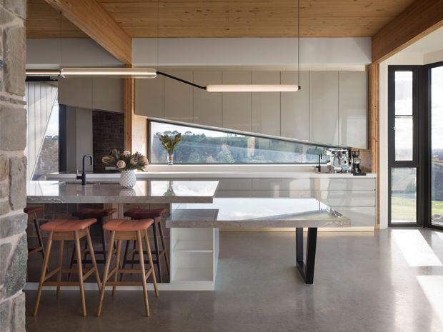 A contemporary open-plan kitchen with a large white island and integrated breakfast bar, timber stools, pale cabinetry, and a long horizontal window overlooking the landscape. The room features exposed timber ceiling beams and boards, a polished concrete floor, stone accents at the left, minimalist pendant lighting, and extensive glazing.