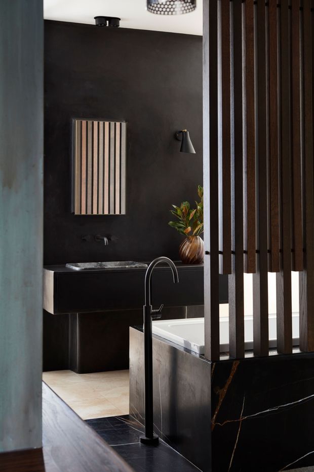 A moody, sophisticated bathroom featuring a freestanding bathtub partially screened by vertical dark timber slats. The room has charcoal walls, a floating dark vanity with a stone basin, black freestanding tapware, pale stone flooring, and a dramatic dark stone bath surround with light veining. A striped wall artwork, sculptural wall light, and vase with foliage add decorative detail.