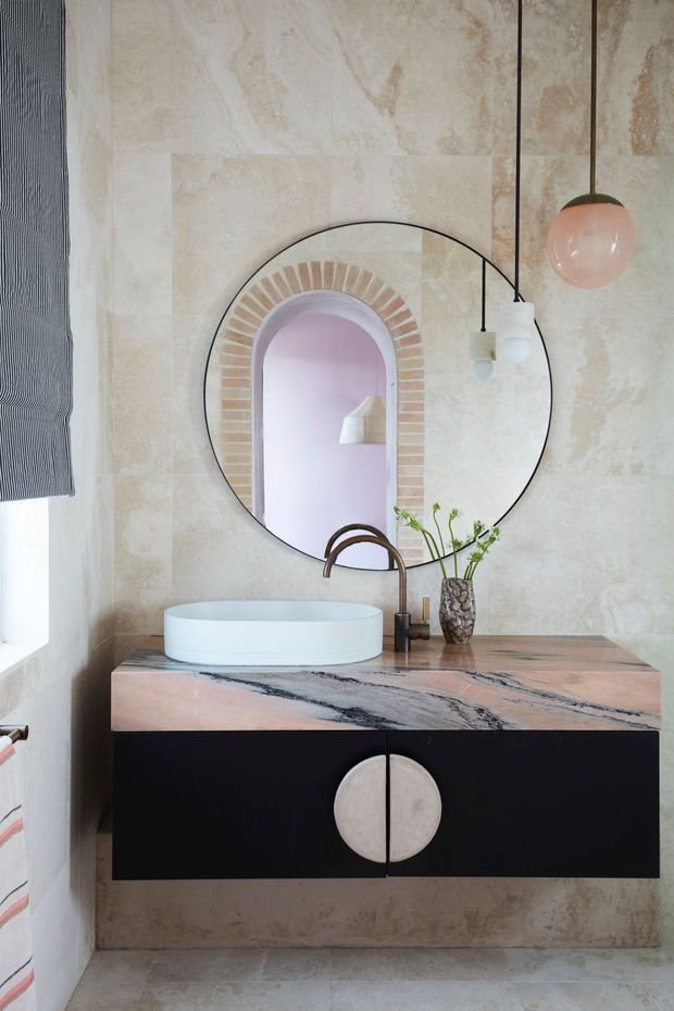 A stylish compact bathroom vanity with a round vessel basin, dark wall-mounted cabinetry, and a pink-veined stone benchtop. The walls are finished in large warm beige stone-look slabs, complemented by a large round mirror reflecting an arched opening with decorative brick detailing. A bronze-toned curved tap, globe pendant light, and small vessel with greenery add warmth and character.