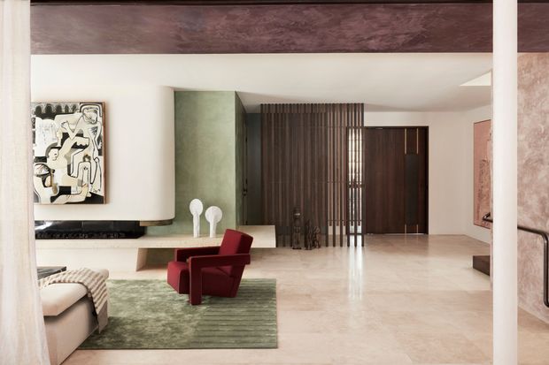 A refined contemporary living room and entry area with a sculptural curved fireplace wall, pale stone flooring, muted green feature wall, dark timber slatted screen and matching timber door. A deep red lounge chair sits on a green rug beside a neutral sofa, while abstract artwork and integrated joinery add visual interest. The space is open, polished and softly lit, with a strong horizontal dark ceiling or beam element.
