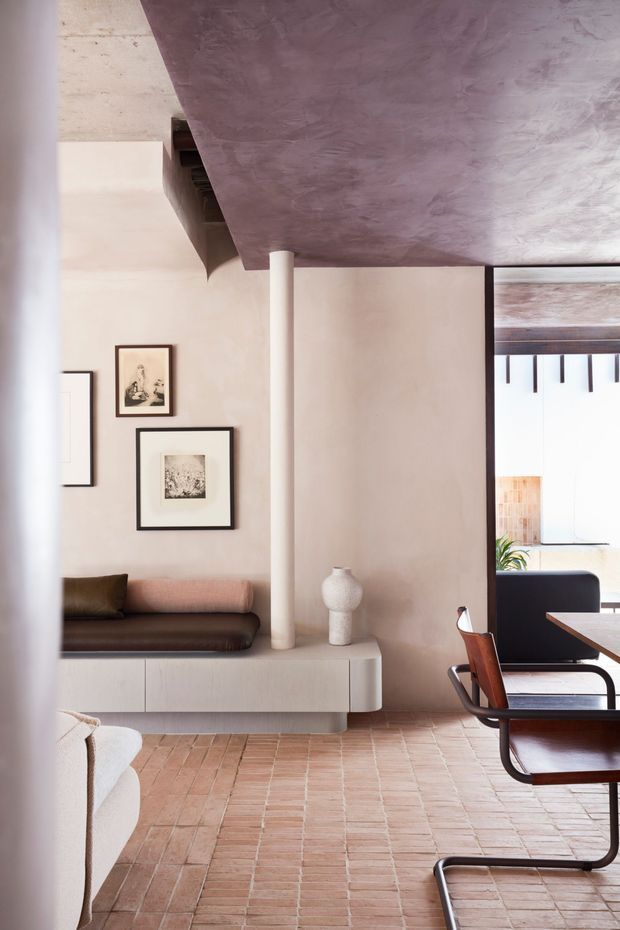 A refined open-plan living and dining interior with a built-in low media or storage console, brown leather seating, framed artwork, and a dining table with tubular-frame chairs. The room features a warm pale plaster palette, terracotta brick flooring, a sculptural mauve-toned soffit or ceiling plane, dark timber detailing, and strong indoor-outdoor glazing. The overall composition is minimalist and contemporary with soft, earthy tones and carefully integrated joinery.