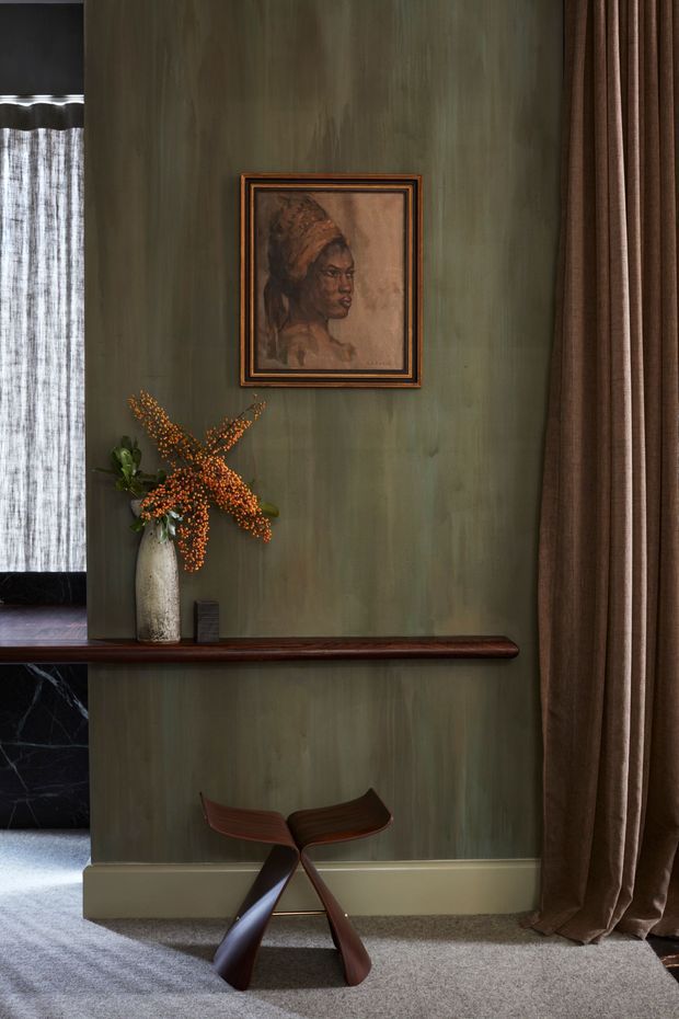 A moody, richly layered interior vignette featuring a deep olive-green painted wall with subtle vertical texture, a framed portrait, a dark timber picture ledge, and a sculptural dark wooden butterfly chair. Floor-to-ceiling brown drapery frames the right side, while a tall textured vase with orange branches adds a warm accent. A glimpse of a window with sheer curtains and dark marble-like wall finish is visible at left.