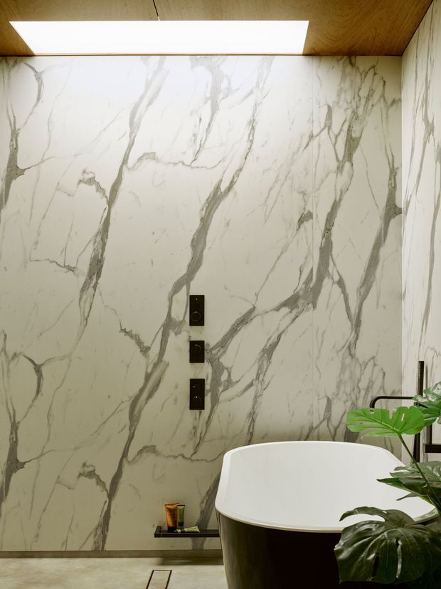 A luxurious bathroom featuring large-format white marble-look wall slabs with dramatic grey veining, a freestanding white bathtub with a dark exterior, black wall controls and tapware, a timber-lined ceiling with a rectangular skylight, and lush tropical plants in the foreground.