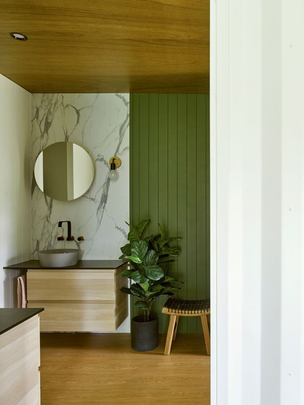 A refined bathroom viewed through a doorway, featuring a floating timber vanity with a dark benchtop, white vessel basin, round wall mirror, black and brass tapware, and a dramatic white marble-look wall finish. Vertical olive-green wall panelling, timber flooring and ceiling, a leafy potted plant, and a small woven stool add warmth and texture.