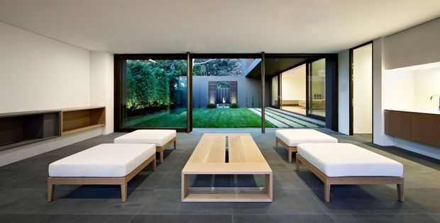 Contemporary living room with a minimalist built-in media wall, low upholstered timber-framed ottomans, and a rectangular timber coffee table. Large dark-framed glazed doors open the room to a landscaped courtyard, while a floating timber console is visible along the right wall. The space has a restrained neutral palette, clean horizontal lines, and dark large-format floor tiles.