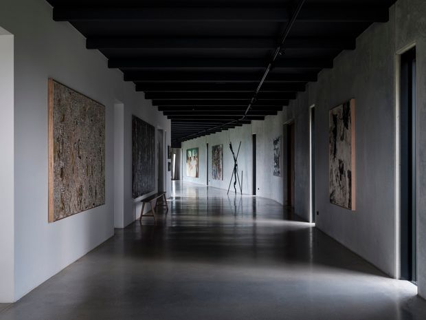 A long, dimly lit gallery-like corridor with polished dark concrete flooring, pale plaster walls, exposed black ceiling beams, and large abstract artworks mounted along both sides. Narrow openings and strong linear perspective lead toward a brighter end, creating a dramatic, minimalist atmosphere.