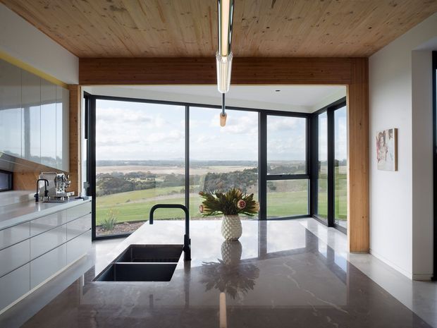 A contemporary kitchen with a large polished stone island, integrated double sink and black gooseneck tap. Full-height black-framed glazing opens to expansive rural views, while a timber-lined ceiling and timber portal frame add warmth. Minimal white cabinetry runs along the left wall, and a linear pendant light hangs above the island.