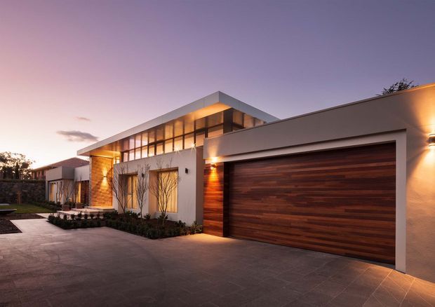 Contemporary single-storey house facade at dusk, featuring a broad flat roofline, extensive upper-level glazing, warm exterior lighting, textured light rendered walls, timber wall accents, and a large horizontal timber garage door. A paved driveway and landscaped planting frame the front entry.