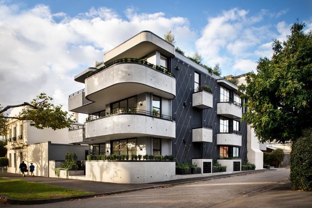 Exterior photograph of a contemporary multi-storey apartment building on a suburban street. The building features rounded projecting balconies with concrete balustrades, dark cladding, extensive glazing, white structural elements, rooftop planting, and a clean modern facade composition.