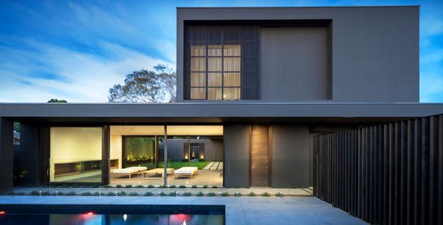 A dusk exterior photograph of a refined two-storey contemporary residence. The home has a strong horizontal composition, dark projecting roof or canopy, smooth dark grey rendered walls, extensive ground-floor glazing, and an upper-level recessed window with vertical screening. A covered outdoor entertaining area and swimming pool are visible in the foreground, with a dark vertical-slat privacy screen at right.