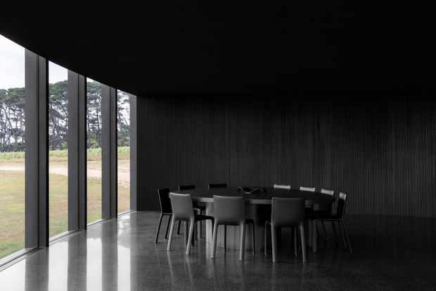 A dark, minimalist dining room with a long rectangular dining table and contemporary chairs arranged beside floor-to-ceiling glazing. The room features a black ribbed feature wall and ceiling, reflective polished dark flooring, and a curved glazed frontage opening toward a grassy landscape.