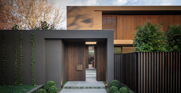 A contemporary residential front entrance viewed from the street, featuring a dark rendered boundary wall, recessed timber pivot door, pale stone-paved path, vertical black metal fencing, timber-clad upper facade with screened windows, and carefully trimmed greenery. The composition emphasizes layered privacy, strong horizontal and vertical lines, and a refined modern entry sequence.