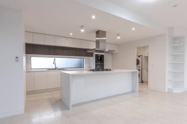 Bright, contemporary open-plan kitchen with a large white island, extensive handleless cabinetry, integrated appliances, a dark timber-look backsplash, stainless-steel rangehood, and pale polished flooring. A laundry area is visible through the doorway at the rear.