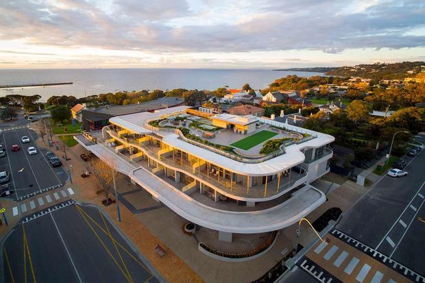Aerial view of a large contemporary coastal mixed-use or commercial building with rounded corners, extensive glazing, broad terraces, and a landscaped rooftop with seating and greenery. The building sits beside a waterfront promenade and urban streets, with low-rise residential development extending along the shoreline in the background.