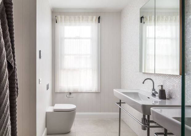 Bright, compact contemporary bathroom with a wall-mounted white basin vanity, freestanding toilet, large mirrored cabinet, pale tiled floor, textured light wall finish, and a window dressed with sheer curtains. Chrome tapware and slim black accents add contrast.