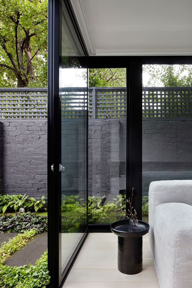 A refined interior corner opening onto a compact planted courtyard through full-height black-framed glazing and a glass door. The space has pale timber flooring, a light upholstered sofa, and a small black cylindrical side table. Outside, a dark painted brick wall topped with a perforated breeze-block screen encloses layered greenery. The composition emphasizes indoor–outdoor connection, strong black framing, and a calm contemporary palette.
