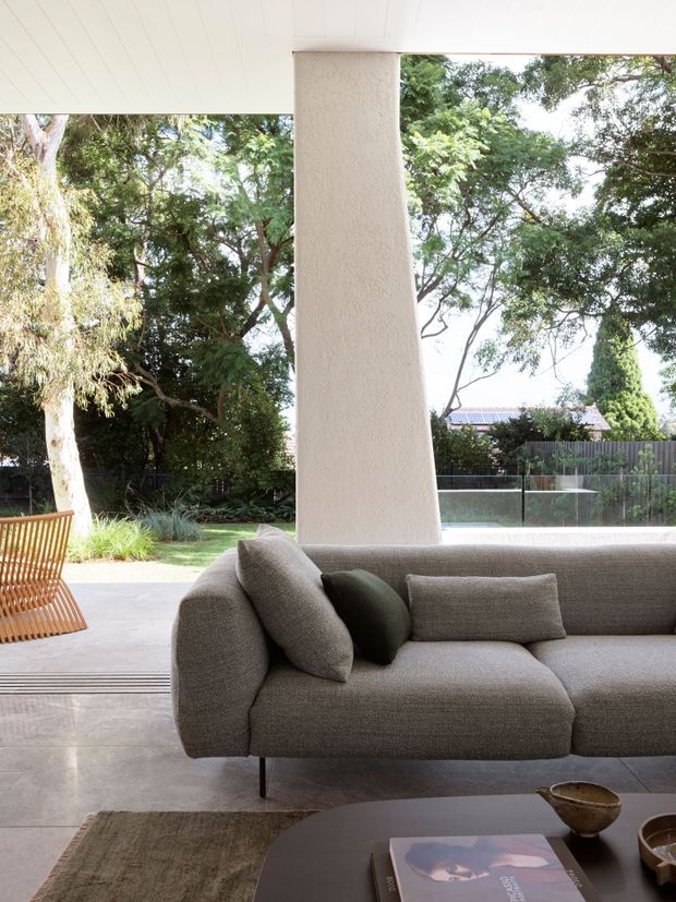 A bright, open-plan living room with a large neutral upholstered sectional sofa in the foreground, set against a broad glazed opening overlooking a leafy landscaped garden and pool area. A textured plaster column divides the opening, while the white lined ceiling and polished concrete floor create a clean, contemporary indoor-outdoor connection. A timber lounge chair, low dark coffee table, rug, and muted cushions add understated warmth.