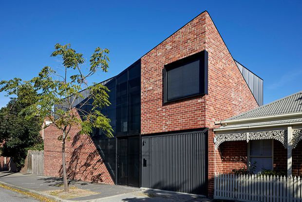 Street-facing view of a contemporary multi-level brick building integrated beside a traditional heritage-style brick house. The new structure has a sharply angled gable-like brick facade, large dark-framed glazing, black vertical corrugated metal garage or entry doors, and dark metal cladding. The adjacent house features ornate lacework verandah detailing, a corrugated pitched roof, and a picket fence. Clear blue sky and street trees frame the architectural composition.