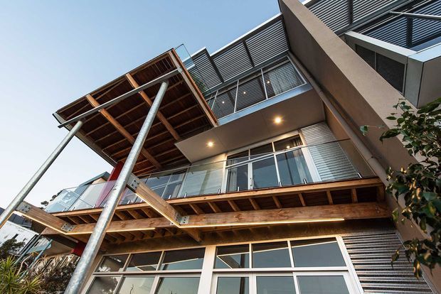 Upward-looking exterior view of a contemporary multi-storey residential building with layered timber balconies, slender metal posts, extensive glazing, horizontal louvres, and projecting roof-like balcony structures. Warm recessed lighting and exposed timber soffits add contrast to the grey rendered facade and aluminium-framed windows.
