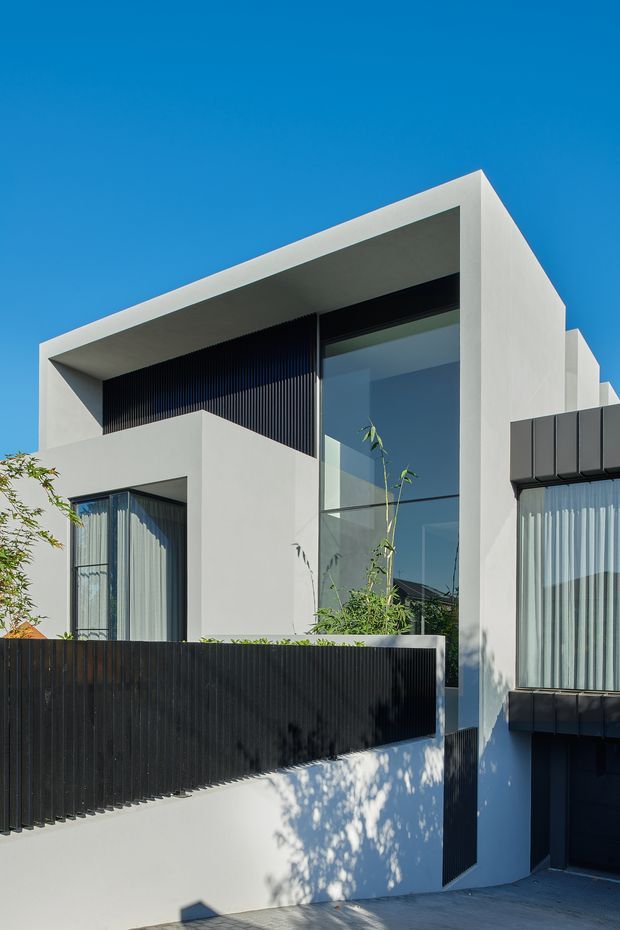 Exterior photograph of a contemporary multi-level residence with crisp white rendered volumes, a strong projecting flat roof, expansive floor-to-ceiling glazing, and dark vertical slatted screens and boundary fencing. The facade is photographed from the street under a clear blue sky, with a driveway and small landscaped planting visible at the base.