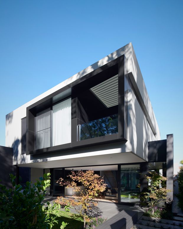 A modern two-storey residence viewed from the garden, featuring crisp white and charcoal geometric volumes, a projecting upper-level balcony or recessed terrace, extensive dark-framed glazing, and a sheltered ground-floor outdoor living area. Lush planting and paved paths surround the house under a clear blue sky.