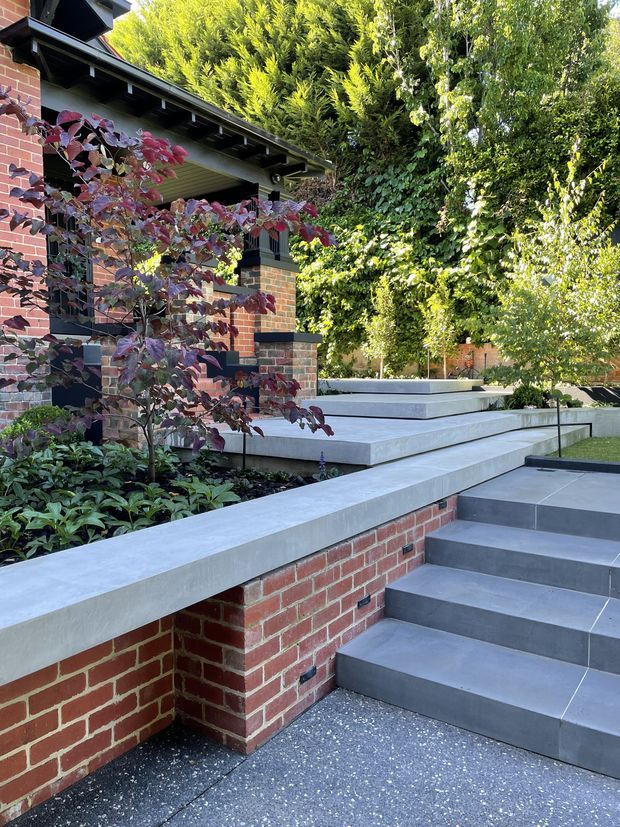 Exterior view of a landscaped residential entry and terrace, featuring red brick walls, dark painted eaves and trim, broad pale concrete coping and steps, dark grey tiled stair treads, and layered garden planting. The stepped levels create a raised courtyard or outdoor approach around the house.