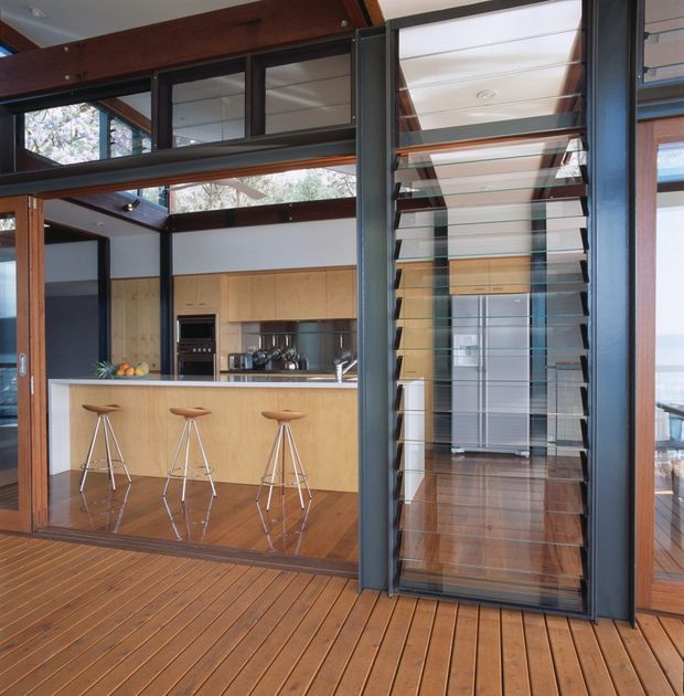 Open-plan modern kitchen viewed from a timber deck, featuring extensive dark-framed glazing, exposed timber beams, a light timber kitchen with island breakfast bar and three stools, stainless-steel appliances, and a prominent glazed louvred screen or door. The space is bright and connected to the outdoors, with warm timber surfaces and strong indoor-outdoor visual flow.