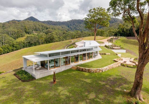 A contemporary single-storey residence set on a gently sloping rural site, surrounded by dense forested hills. The building has a long, low horizontal form with extensive floor-to-ceiling glazing, white flat roof planes, slim framing, and an elevated covered terrace. Curved stone retaining walls and landscaped lawns define the outdoor spaces.