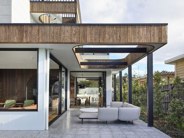 Contemporary multi-level residential home viewed from a covered outdoor terrace. The facade combines white rendered surfaces, dark vertical structural posts, and extensive warm timber cladding. Layered cantilevered balconies and curved-edged overhead openings create a strong modern architectural composition. A large outdoor lounge with a light upholstered sectional sits on grey paved flooring, while glazed openings reveal an interior dining area and living spaces. Landscaped planting and neighbouring residential structures are visible beyond the terrace.