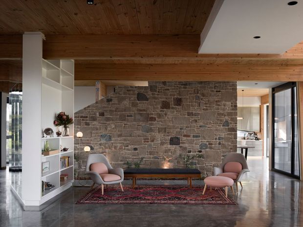 A refined open-plan living room centred on a large rustic stone feature wall with a linear fireplace. The room has a warm timber-lined ceiling, polished concrete flooring, extensive glazing, a built-in white display shelving unit, two pink upholstered lounge chairs, a low timber-and-dark upholstered bench, and a patterned rug. A kitchen is visible beyond the opening on the right.