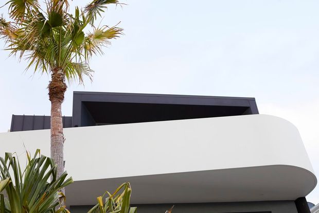 Close-up exterior view of a contemporary building with a crisp white, smoothly curved balcony or overhanging facade element, dark charcoal upper roof or wall planes, and tropical palm planting in the foreground. The composition highlights the rounded balcony edge and contrasting geometric dark roof form against a pale sky.
