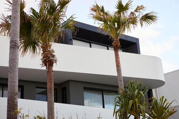 Exterior view of a contemporary multi-level residence featuring a dark charcoal facade, broad white curved balcony parapets, large glazed openings, and tropical palm landscaping.