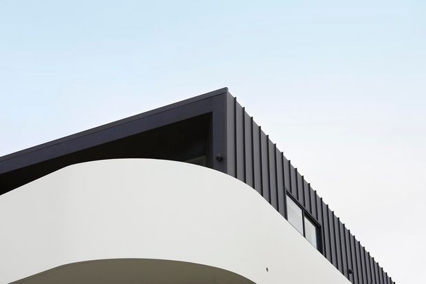 Close-up architectural exterior view of a contemporary building corner, featuring a dark ribbed metal-clad upper facade and a smooth white curved wall or balcony edge beneath it. The composition emphasizes strong geometric lines, layered roofline detailing, and curved forms against a pale sky.