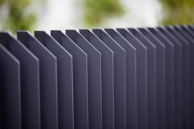 Close-up product-style image of a repeated dark charcoal architectural screen or privacy-fence element, formed from evenly spaced vertical panels with angled tops or folded profiles. The background is softly blurred with greenery, emphasizing the geometric rhythm and matte finish of the structure.