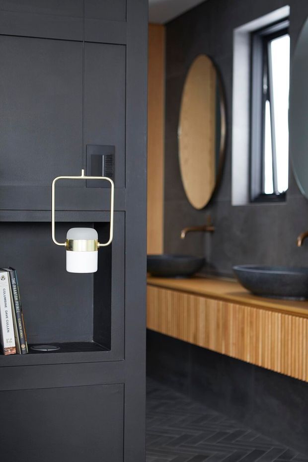 A sophisticated dark-toned bathroom featuring a double vanity with fluted timber cabinetry, a wood benchtop, black vessel basins, and wall-mounted bronze-toned taps. The space includes dark tiled flooring and walls, a round mirror, black-framed window, and integrated charcoal wall panelling with a small decorative pendant light and open shelf.