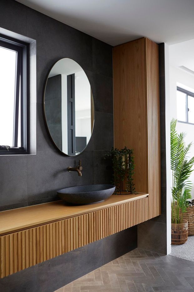 Contemporary bathroom vanity with a floating fluted timber cabinet and matching vertical timber screen detail. A dark stone or porcelain tiled wall provides a dramatic backdrop for a round mirror, black vessel basin, and wall-mounted bronze tapware. The room includes a warm timber benchtop, patterned grey floor tiles, dark window frames, and indoor greenery near the adjoining bright space.