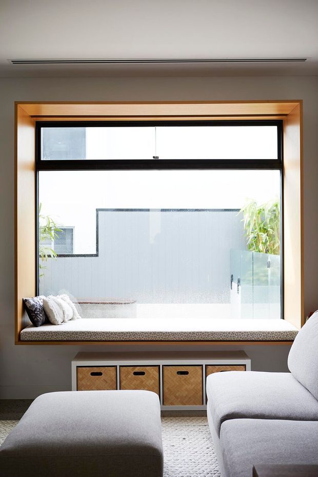 Bright, contemporary living room centered on a large black-framed picture window with a built-in timber-framed window seat and cushions. A low timber storage console sits below the window, while a pale upholstered sofa and ottoman occupy the foreground. The room has a neutral palette, clean lines, and abundant natural light.