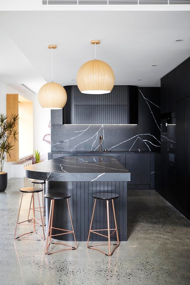 A contemporary kitchen featuring a large island with a curved breakfast-bar extension, dark vertically fluted cabinetry, and dramatic dark stone surfaces with white veining. Three oversized spherical pendant lights hang above the island, while rose-gold metal bar stools provide seating. The space has a polished concrete or terrazzo-like floor, white ceiling, integrated appliances, and a bright adjoining entry or living area with plants.