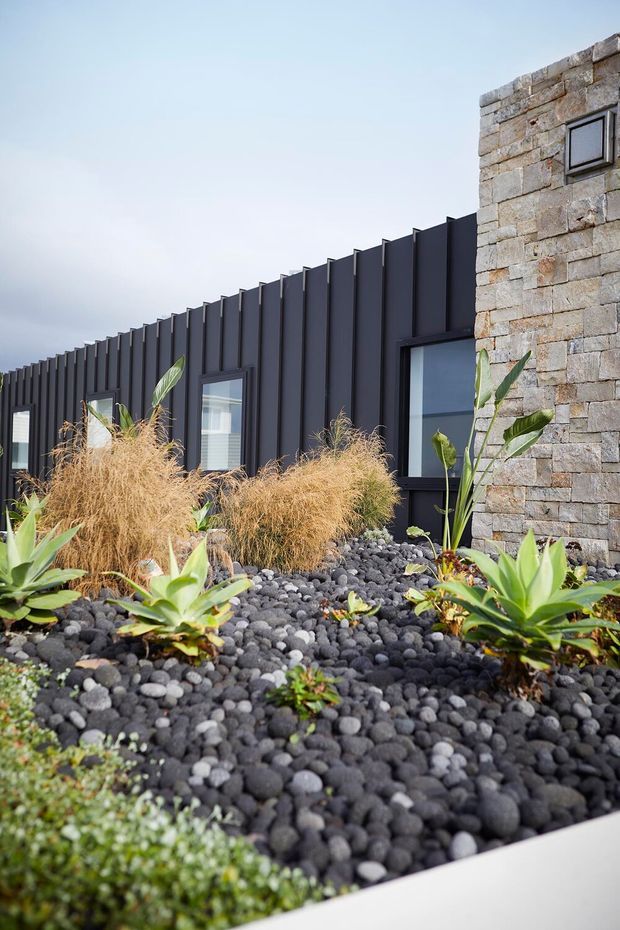 Exterior architectural view of a contemporary building with dark vertical metal cladding, large windows, and a contrasting rough-cut stone wall. The foreground features a landscaped rooftop or raised garden bed covered in dark rounded stones, ornamental grasses, and sculptural green plants.